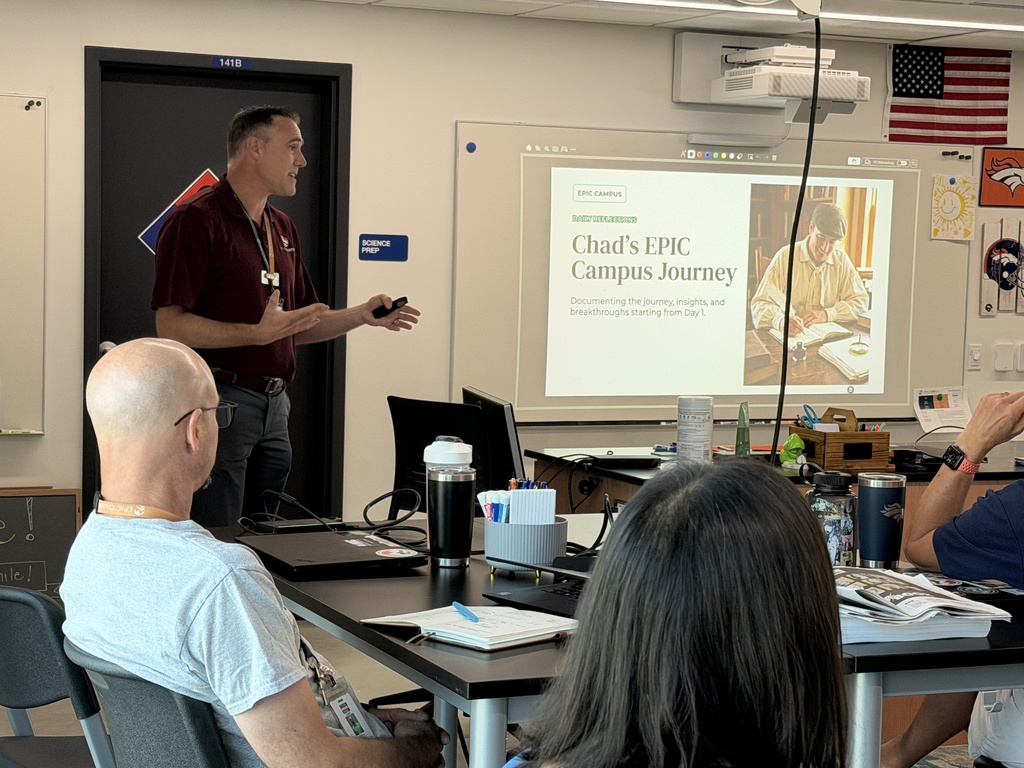 A man in a maroon shirt gives a presentation in a classroom with a whiteboard, projector, and an American flag.