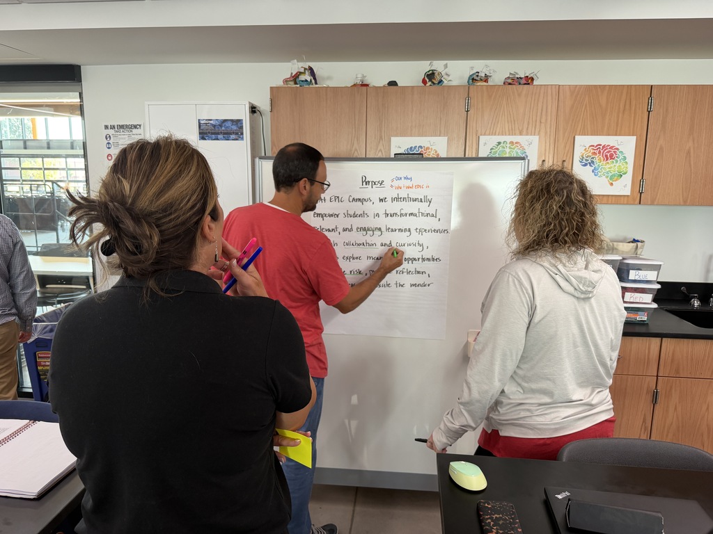 Four people are in a room. A man writes on a whiteboard. One woman watches him write.