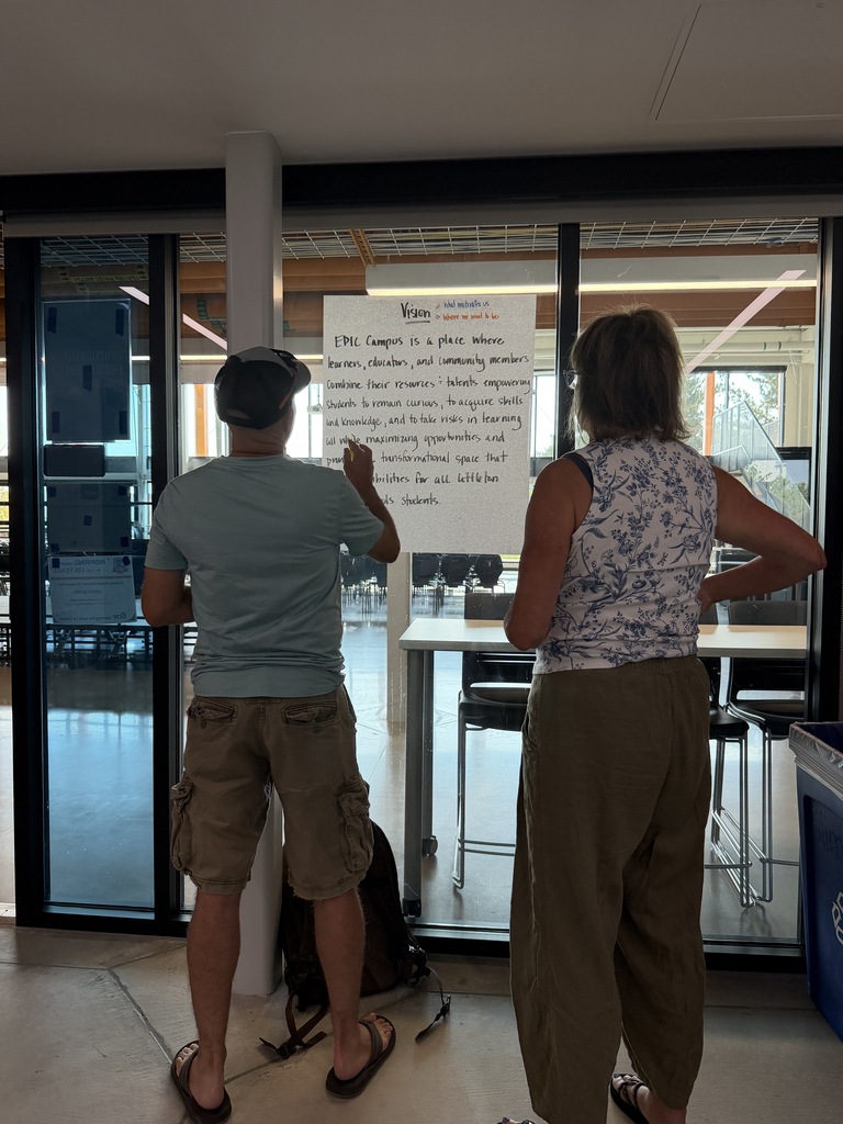 Two people stand by a glass door, with one person writing on a white board with text.