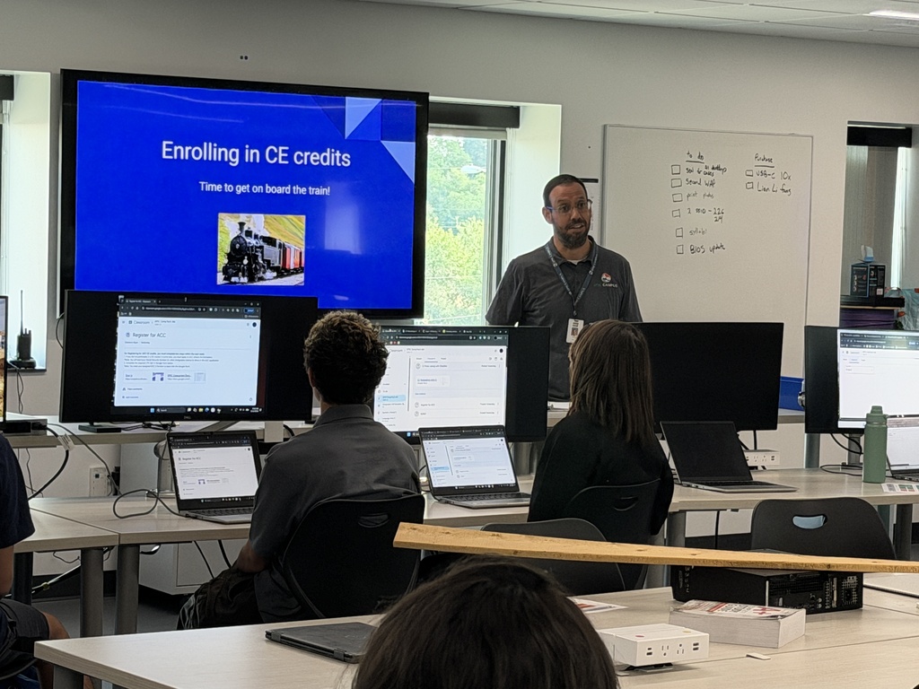 A classroom with a large screen showing "Enrolling in CE credits." A man speaks to others seated at desks.