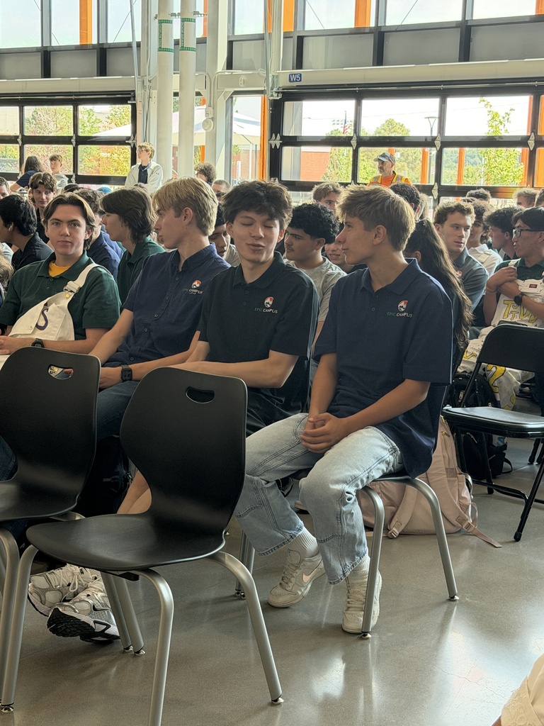 Group of people wearing matching blue shirts seated in chairs in a large room with large windows.