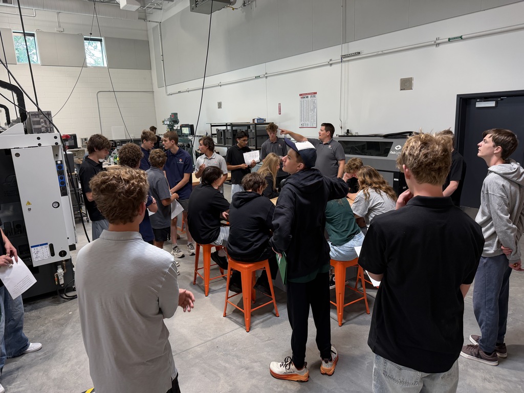 A group of people stand around, listen to a presentation, and view a large machine in a workshop.