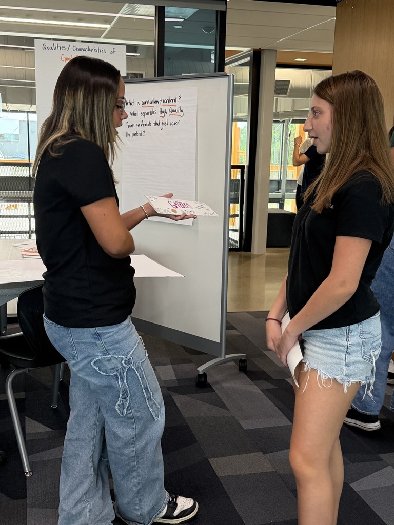 Two women stand facing each other in a room with a whiteboard and open doors. One holds paper.