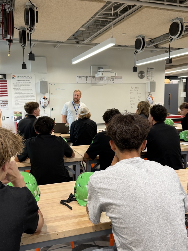 Students in a classroom setting, wearing hard hats, seated around tables, with a teacher standing in front of a whiteboard.