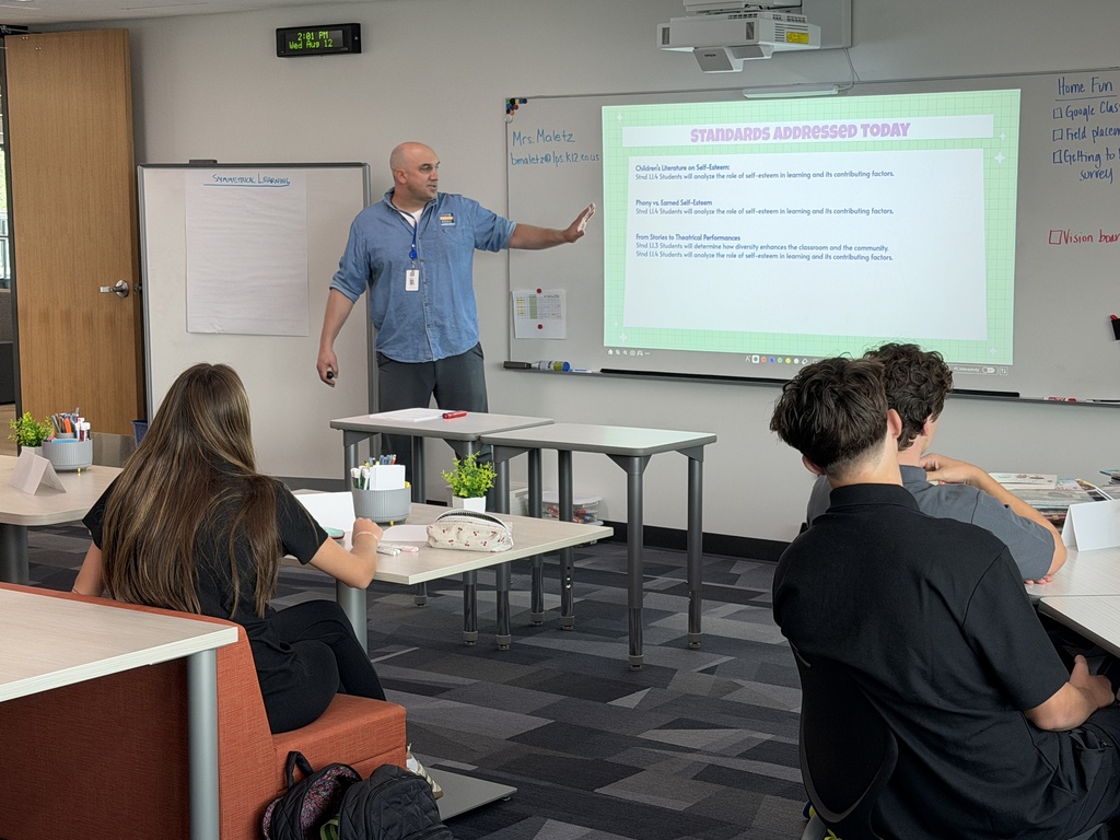 A classroom with a man standing at a podium, presenting a slideshow on a projector screen. Students sit at tables.