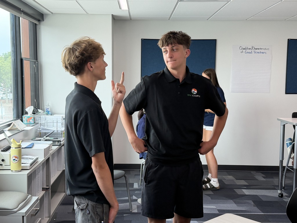 Two men face each other in an indoor office. One man makes a peace sign. There are desks, chairs, and posters on the wall.