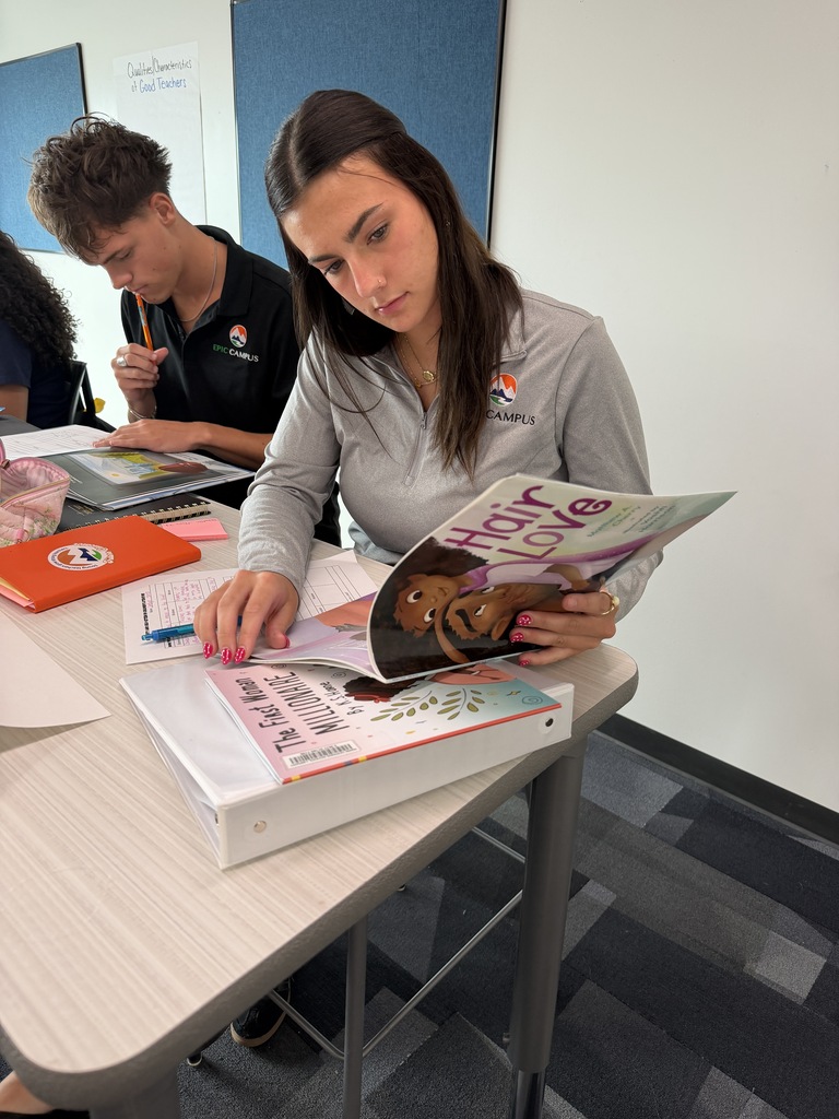 Woman reads a book titled "Hair Love" at a desk. Behind her, a man writes on a piece of paper.