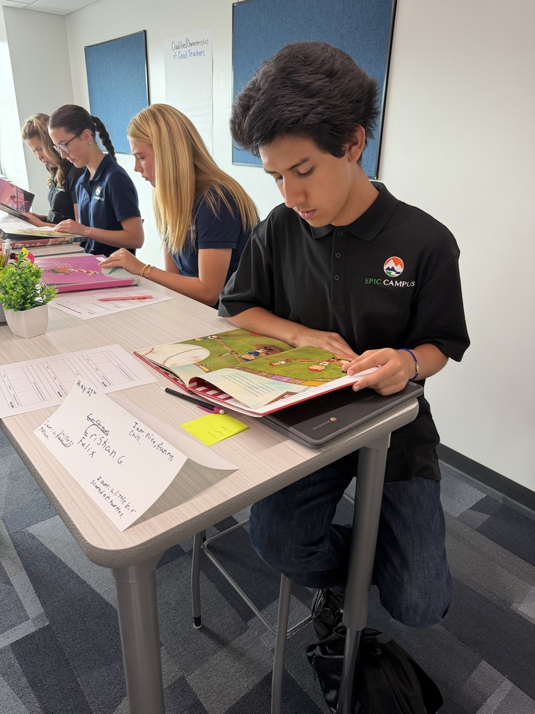 Group of people sitting at a table, a person focused on a book, papers, and a potted plant on the table.