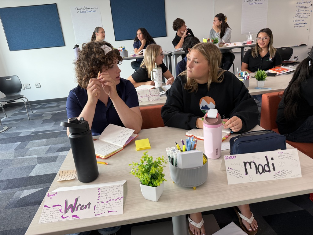 A group of women seated in an indoor classroom, some wearing shoes, engaged in discussion. Tables are cluttered with books, pens, and name tags.