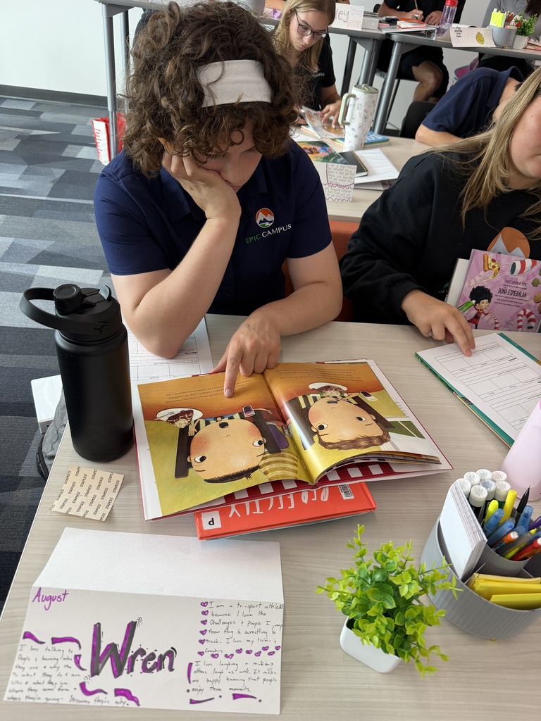A person reads an open book with a picture on it, while another person sits nearby, focused on their work.