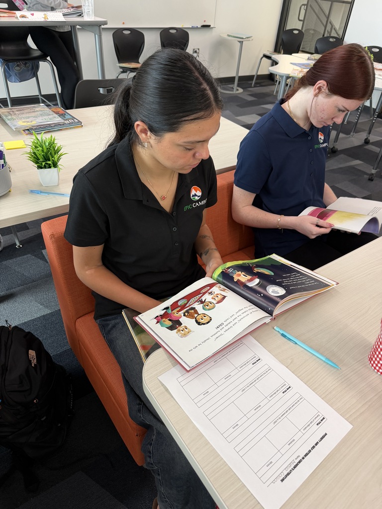 Two women sit in chairs at a table, each with an open book and pen. One has a backpack nearby.