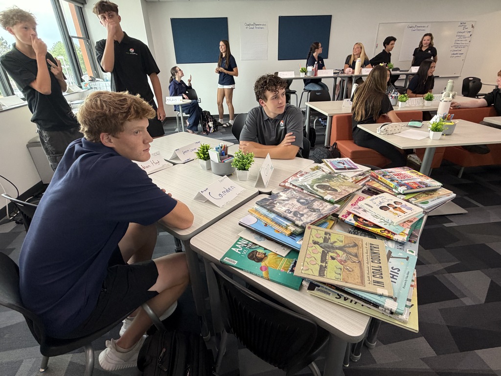 A classroom with students seated at tables. One student sits with books and plants. Others are behind them.