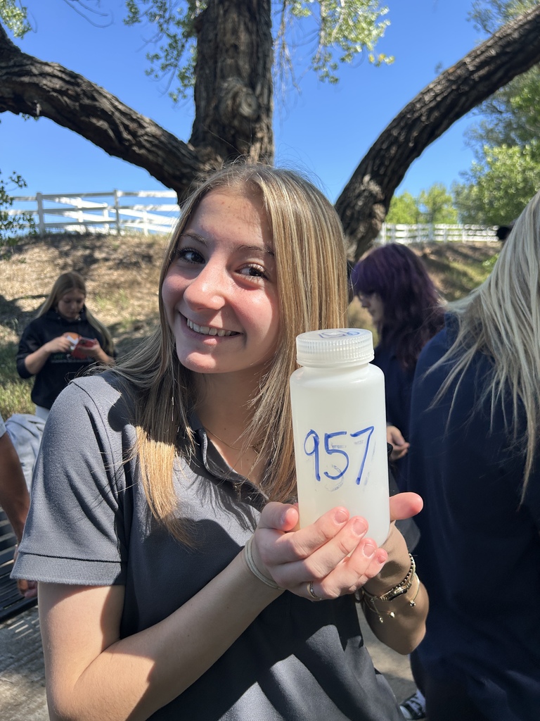 Woman holding a bottle with a number. She has blonde hair. Blurry background of people and trees.