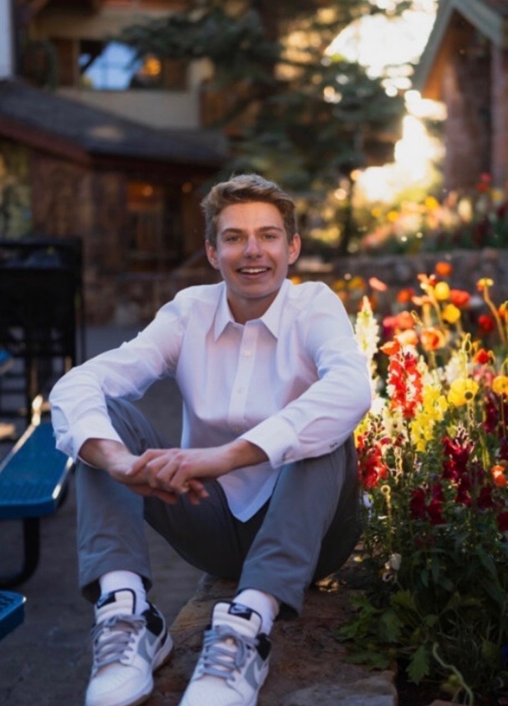 A man sits on a rock with flowers behind him, smiling, in a park. He wears a white shirt and gray pants.