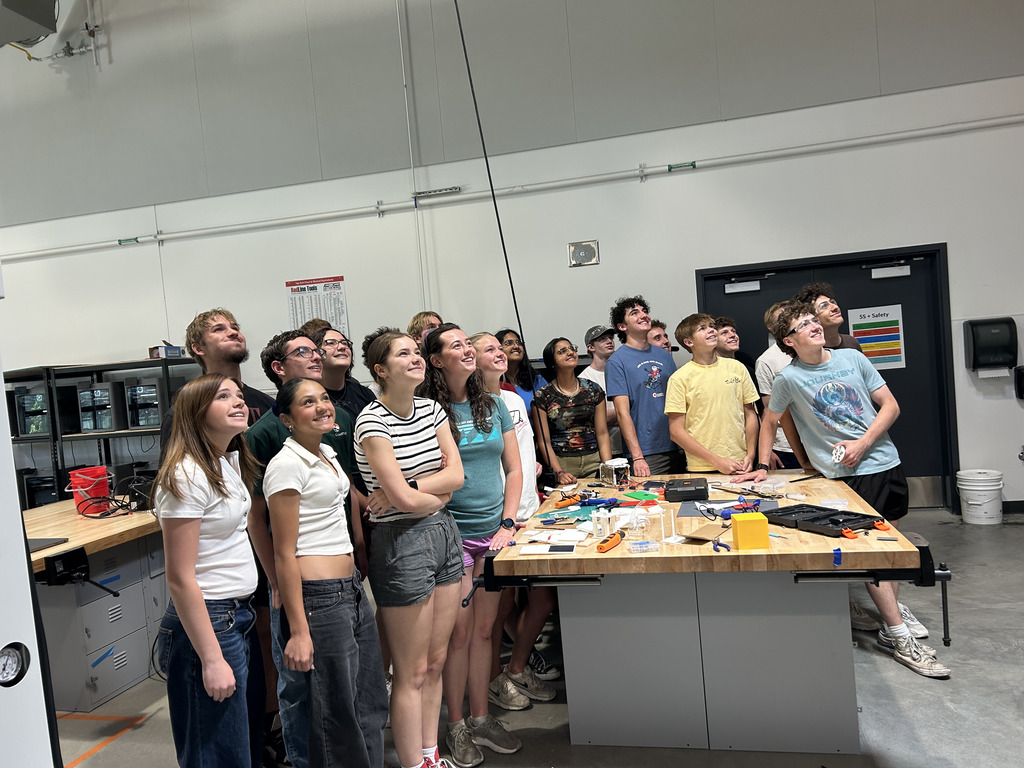 A group of students are standing around a table with various items, looking upwards.