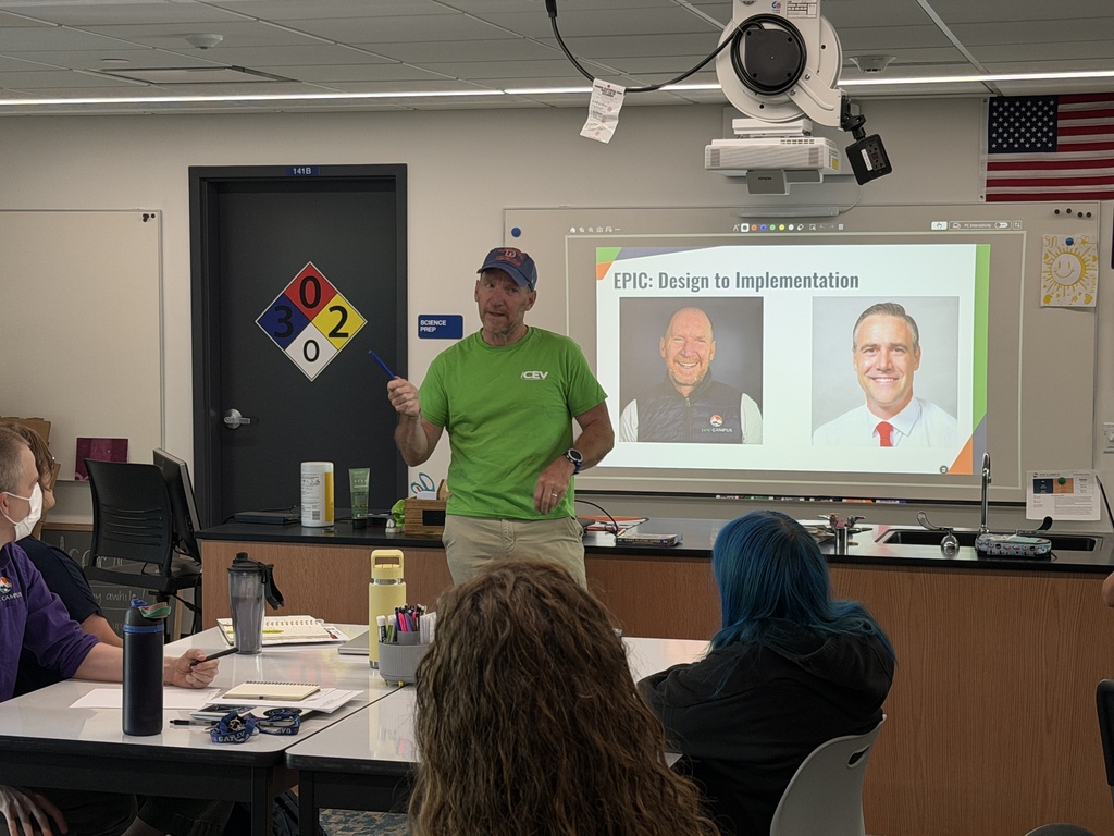 A man stands before a screen in a classroom, speaking to a group. The screen shows two men.