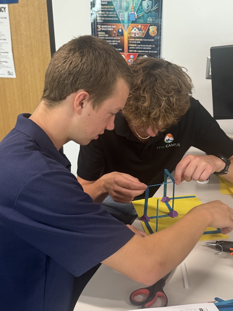 Two people in blue and black shirts assembling a small model with scissors on a table.