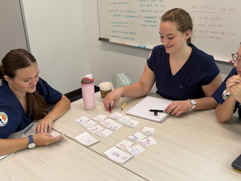 Three individuals in blue uniforms seated at a table with notes, markers, and a cup. One person smiles and gestures to cards on the table.