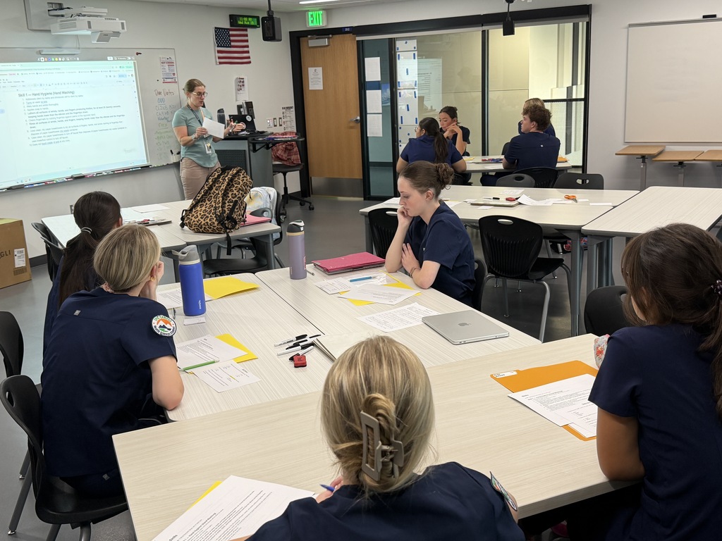 People wearing uniforms sit around a table in a classroom, facing a woman at a front projector screen.
