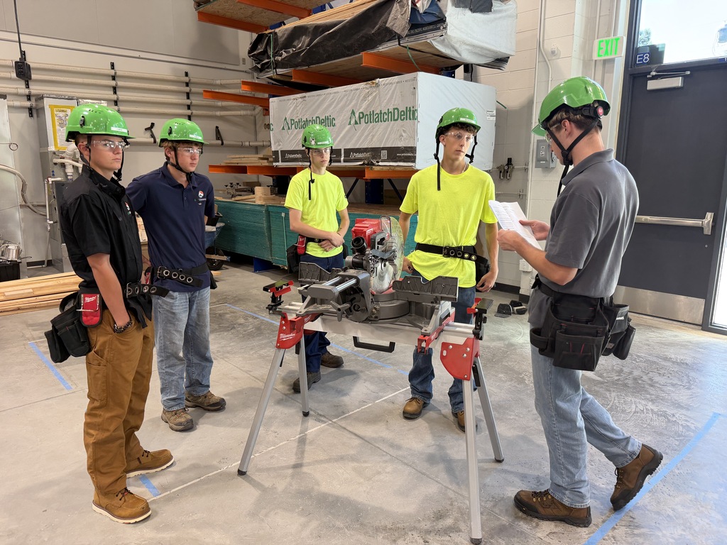 Five men in green helmets and work boots gather around a circular saw in a workshop. One holds paper.