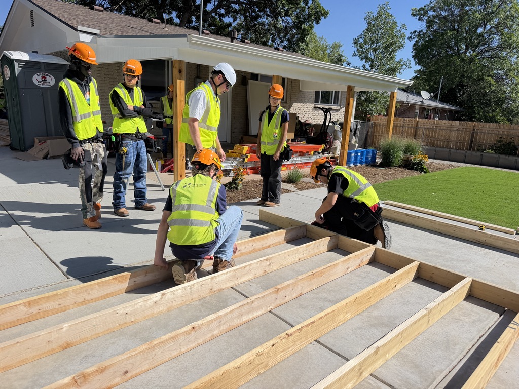 Seven people in bright yellow vests and hard hats are building a deck in a backyard.