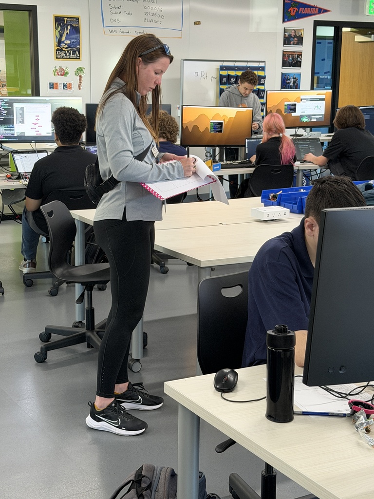 Person in black clothes stands in a classroom with computers on tables, holding paper and looking down.