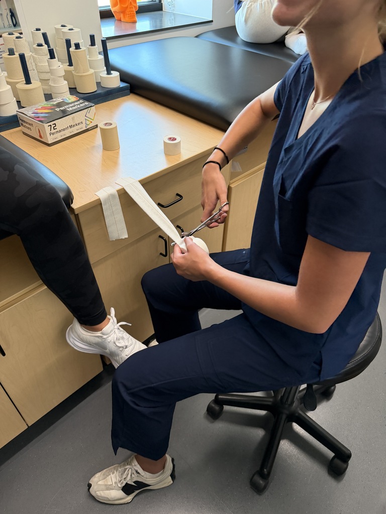 A person in blue scrubs sits on a chair, cutting tape near another person's legs. A wooden desk with a black mat behind them.