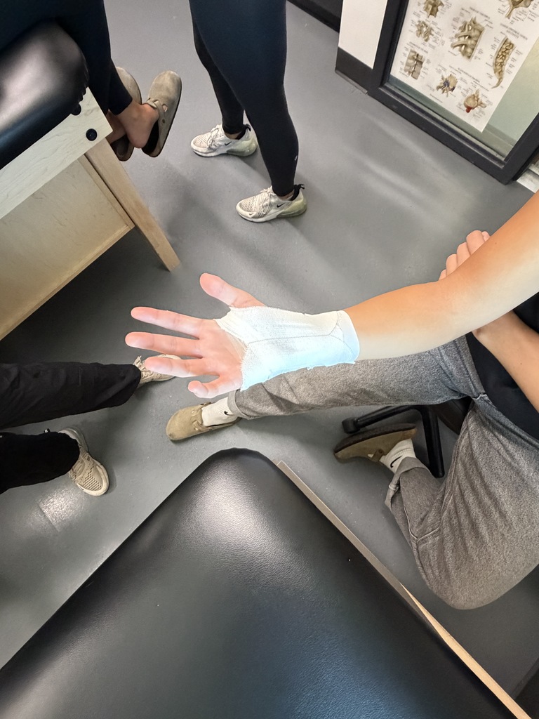 Four individuals are indoors. One has a white bandage on their right wrist, another is sitting with their legs crossed.