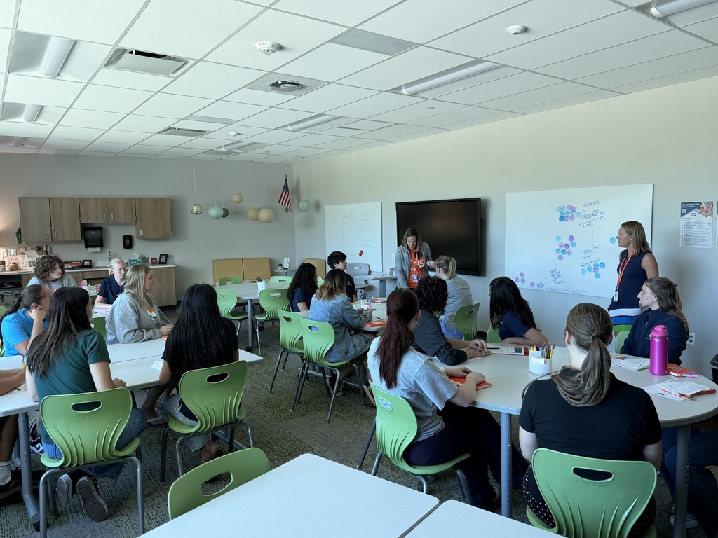 Group in chairs around tables in a classroom. A woman stands at the front by a whiteboard.