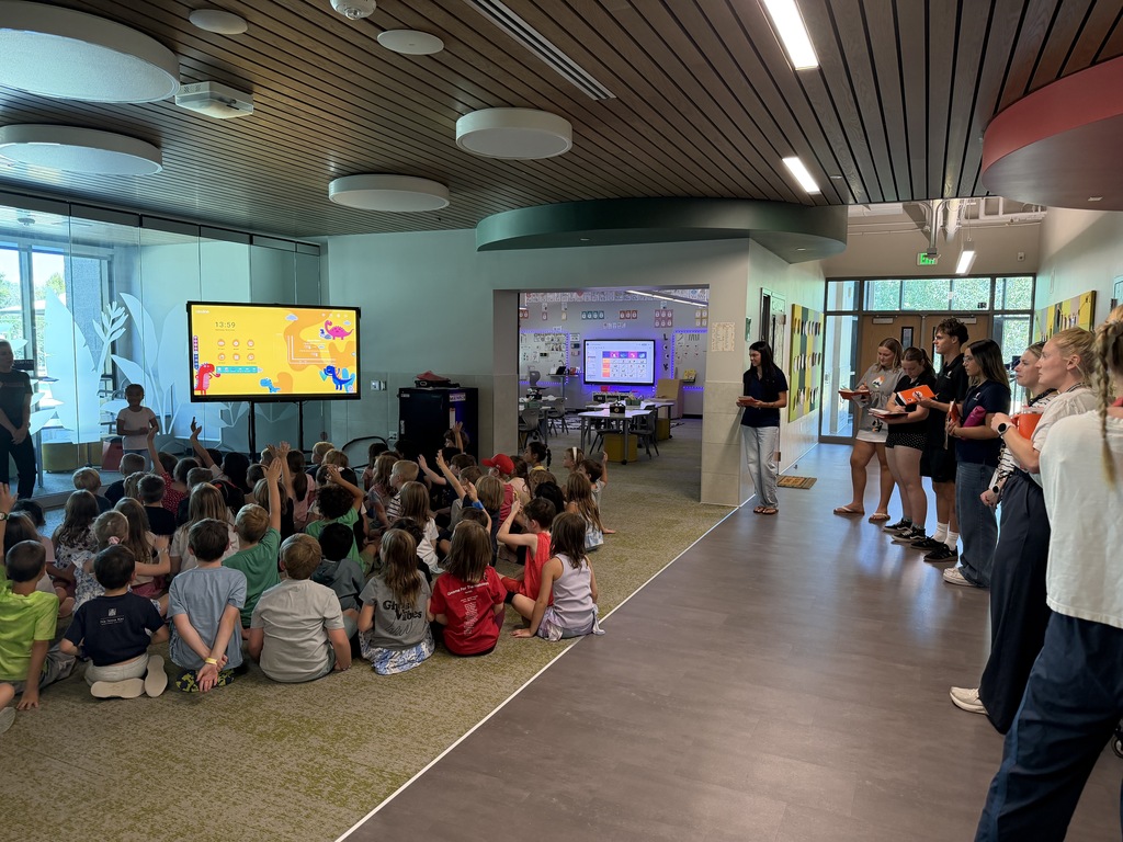 Indoors, a group of children sit in a circle on the floor, listening to a presentation on a large screen.