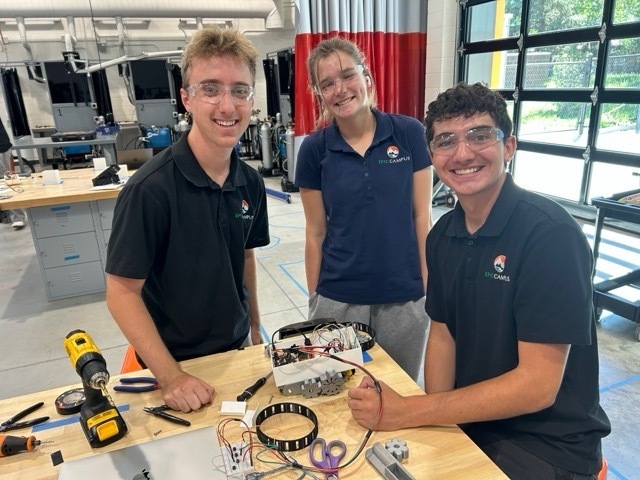 Three young adults in a workshop, surrounded by tools. One holds a drill, another a tool box.