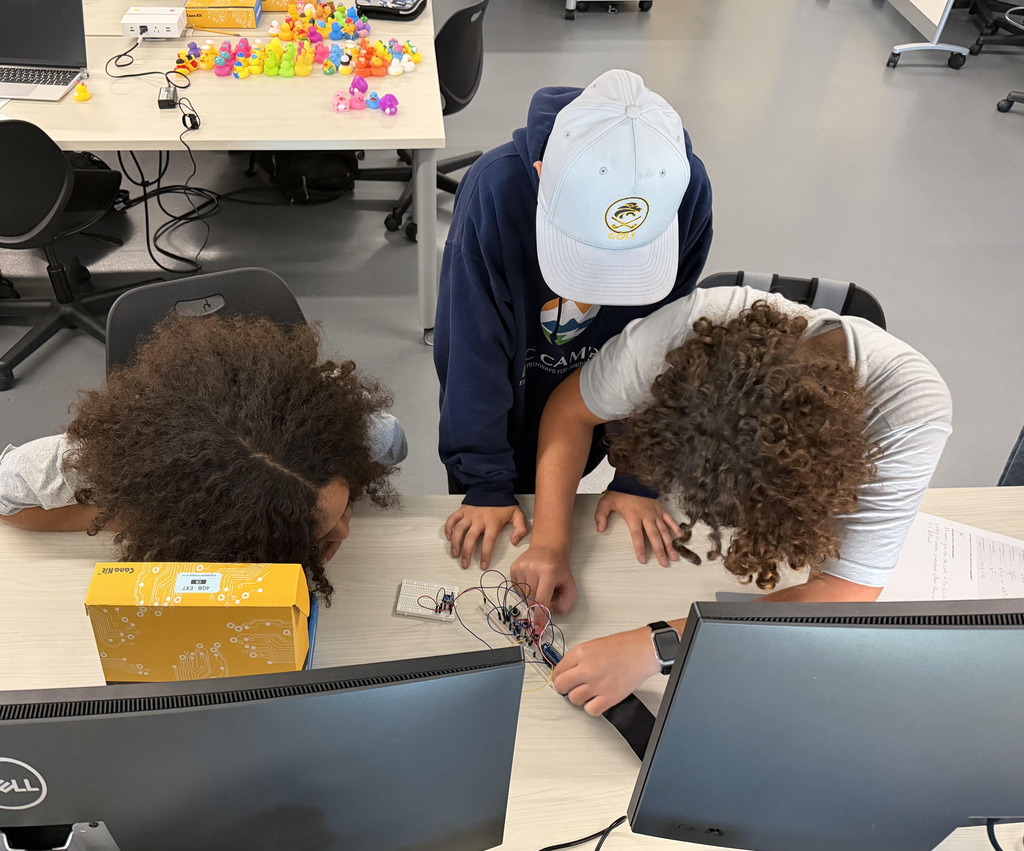 Three people, wearing hats, are seated at a table, working on an electronic project.