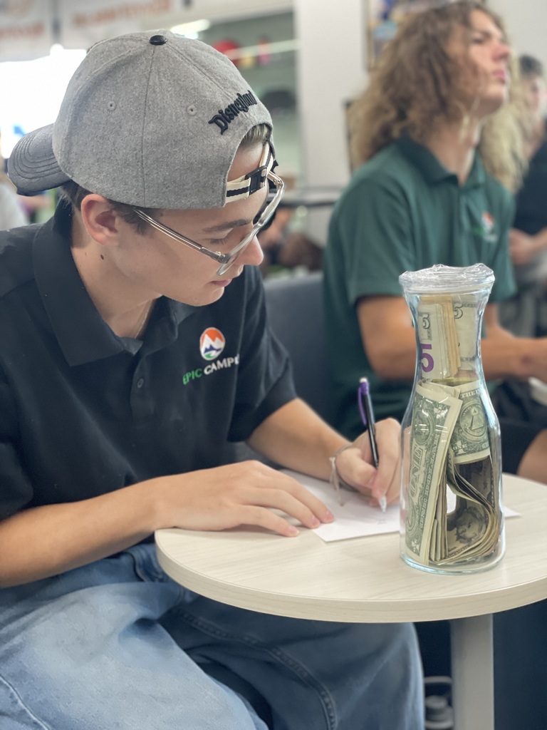 Person wearing a cap and glasses sits at a table, writing on paper with a pen.