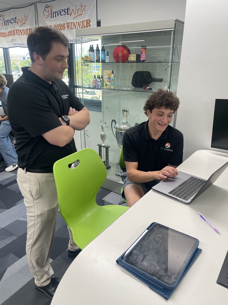 Two people in an office. One person in black shirt and khakis stands with arms crossed. Another person sits with a laptop in front of them on a desk.