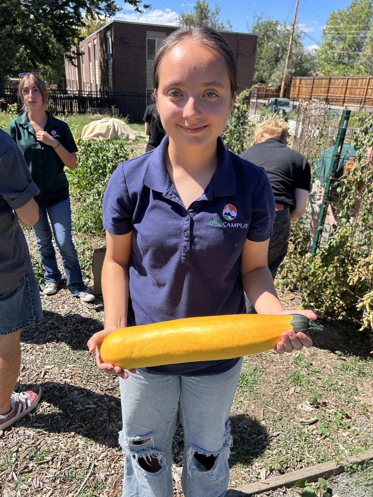 Woman in a blue shirt holds a long, yellow vegetable in a garden with others. Blurry background shows plants and buildings.