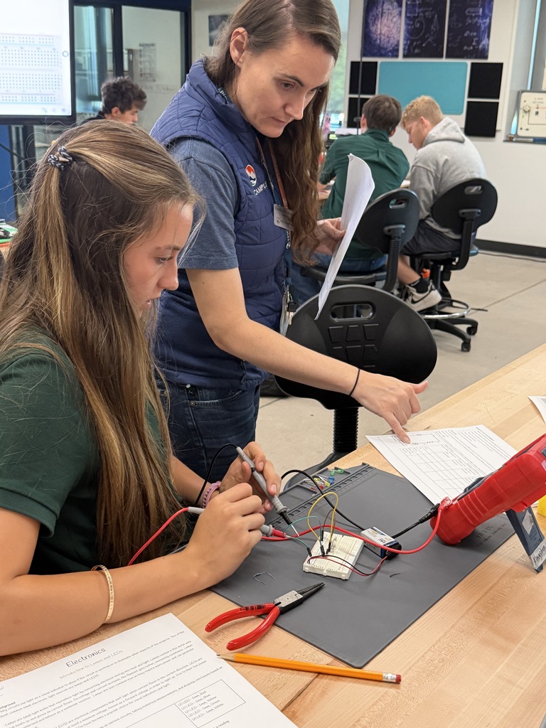 Two women in a lab setting working with soldering irons and wires on a desk. Other students and computers are in the background.