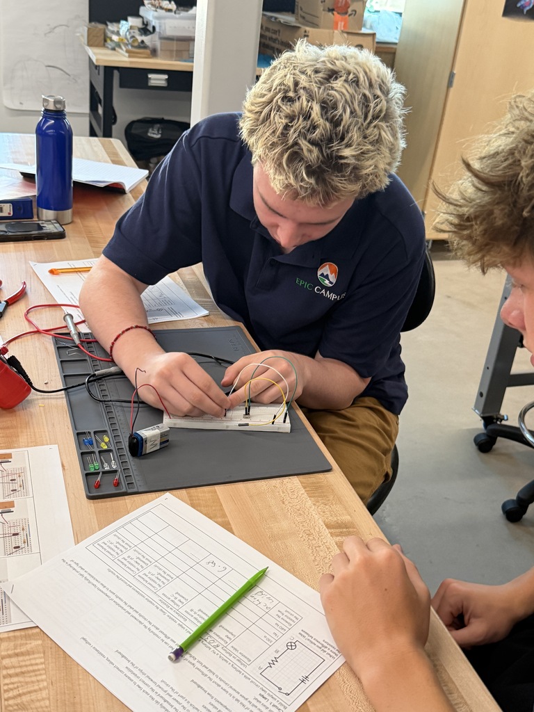 Two individuals, one in a blue shirt, are seated and collaborating on a circuit board in a workshop.
