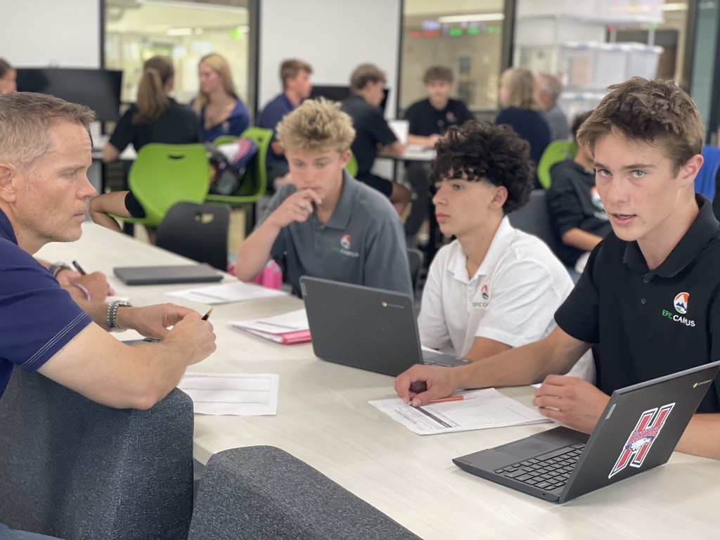 A group of students and an instructor are sitting at a table in a classroom. They have laptops and papers in front of them.