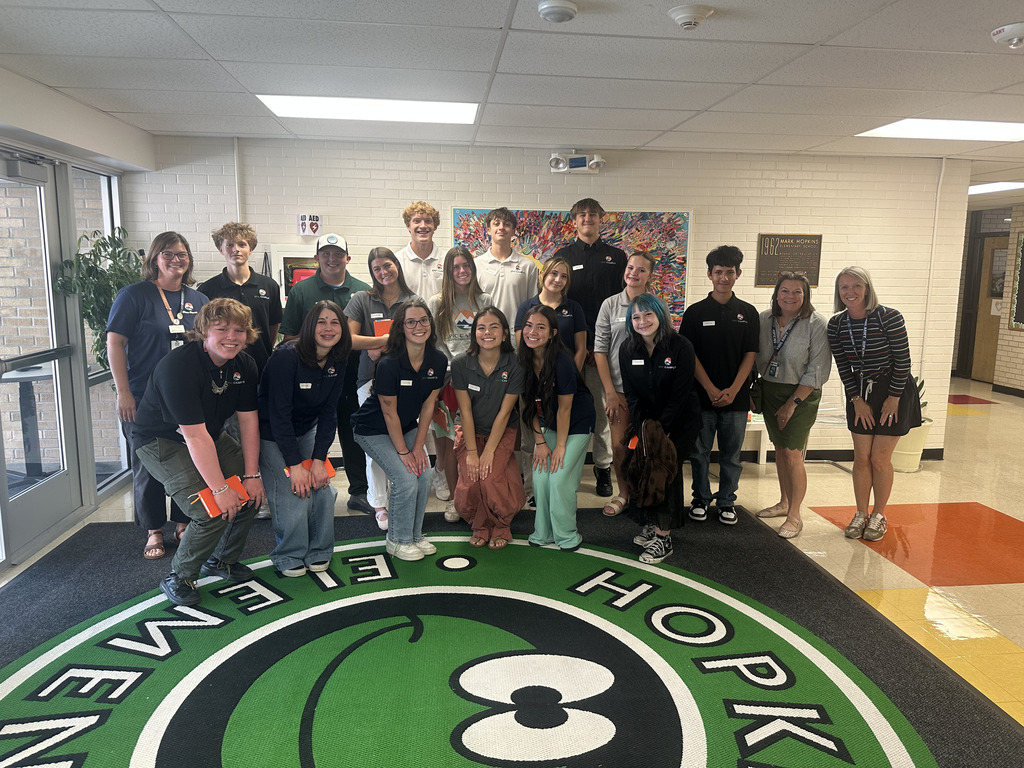 A group of students, faculty, and staff pose in front of a green and white logo on the floor.