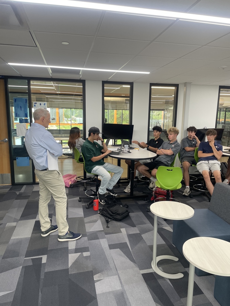 A man stands in a modern room; six people sit around a table, conversing with him.
