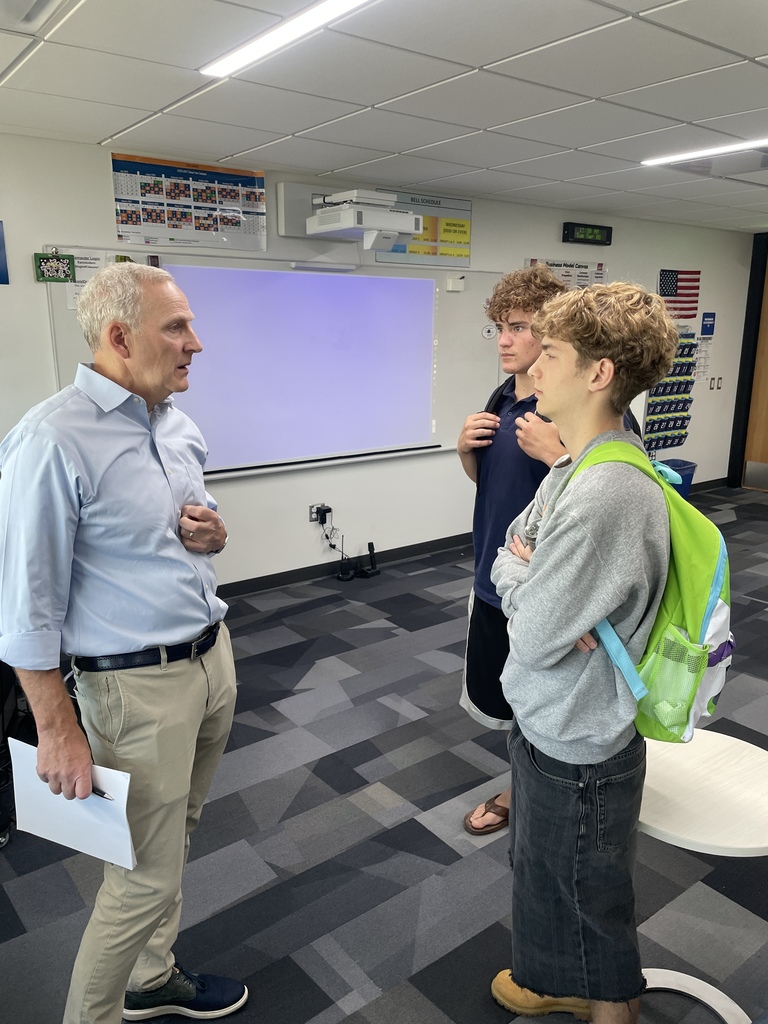 Three people in a classroom. A man in blue speaks to a woman in gray. Behind them, a whiteboard is attached to the wall.