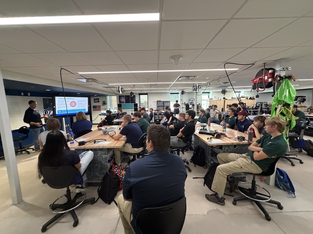 Many people sit at tables in a room, facing a person at a podium. A monitor is on the podium.