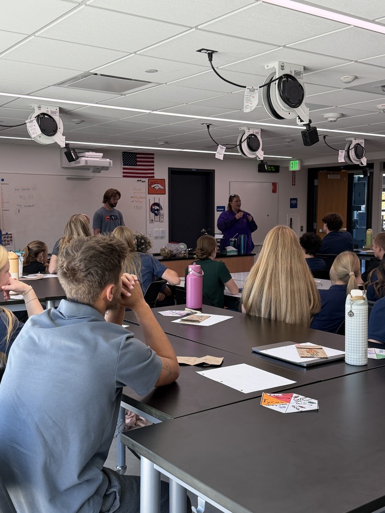 Classroom with students seated at tables. A teacher stands at the front, and a whiteboard is in the background.