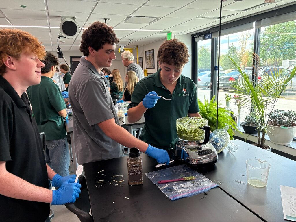 Three students in a lab. One using a food processor. Another holding a spatula. All wearing gloves.