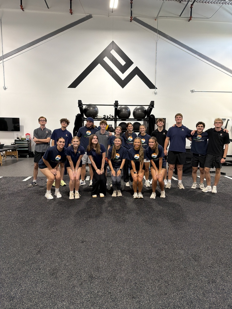 Group in blue shirts and shorts posing for photo in gym with weights and logo on wall.