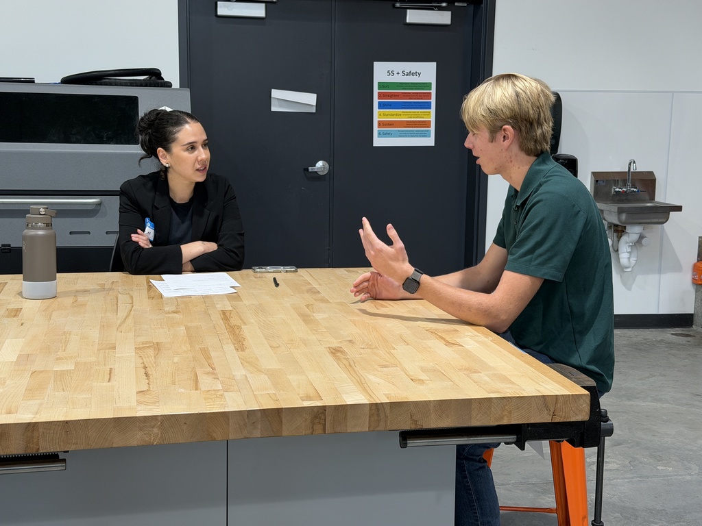 A man and woman seated, engaged in conversation. The man wears a green shirt and wristwatch. Behind them, a gray door with a multicolored chart.