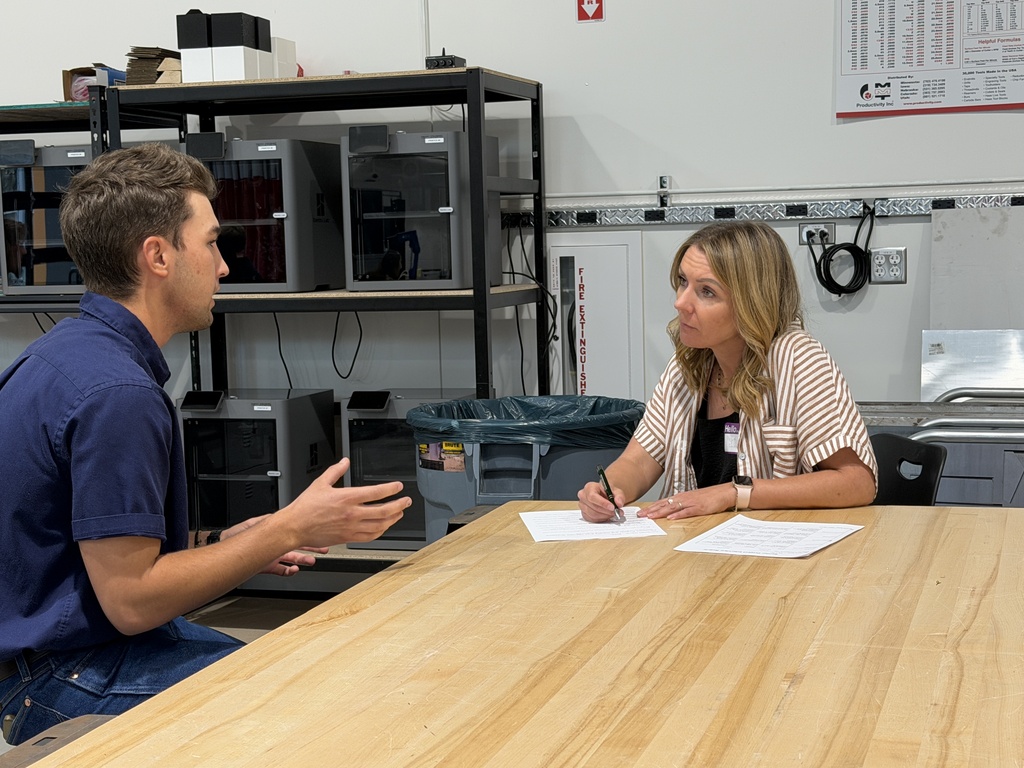 Two people at a table. One person is writing on paper with a pen. The other is talking.