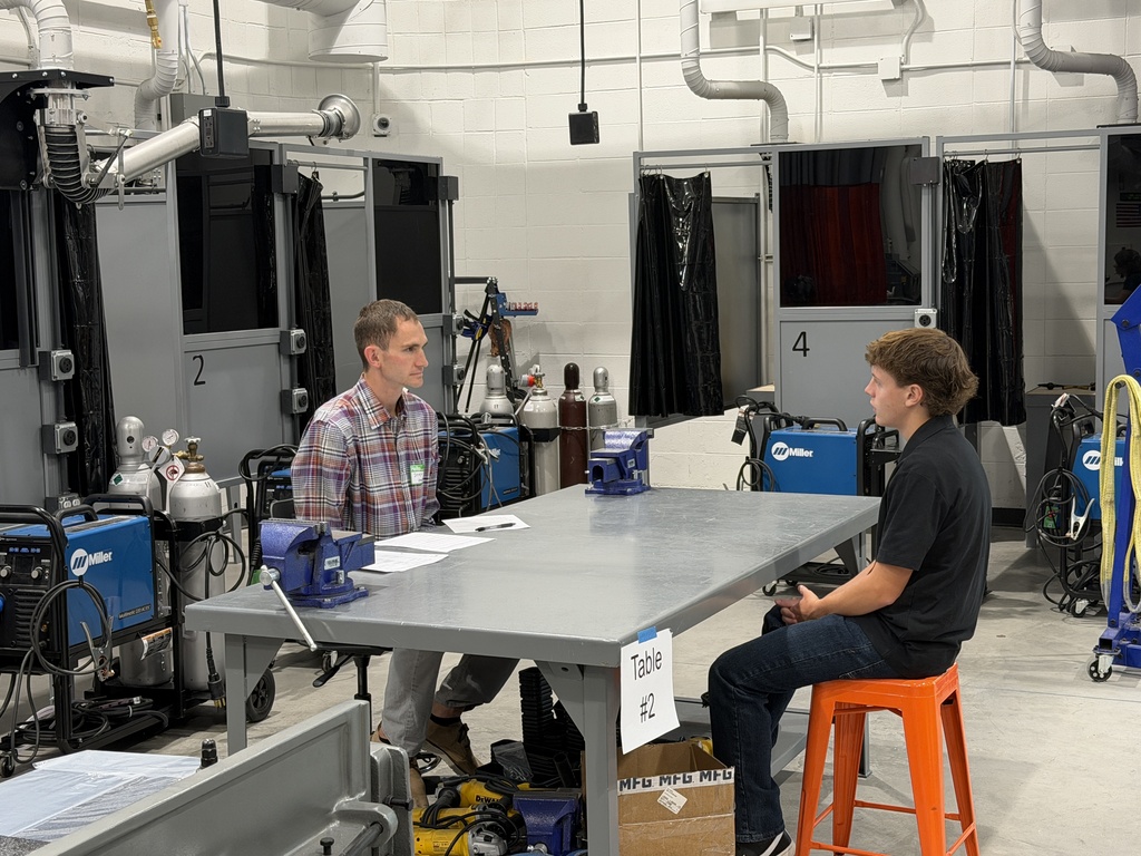 Two people sit at a table in an industrial space with machines. One wears a plaid shirt and the other a black shirt.