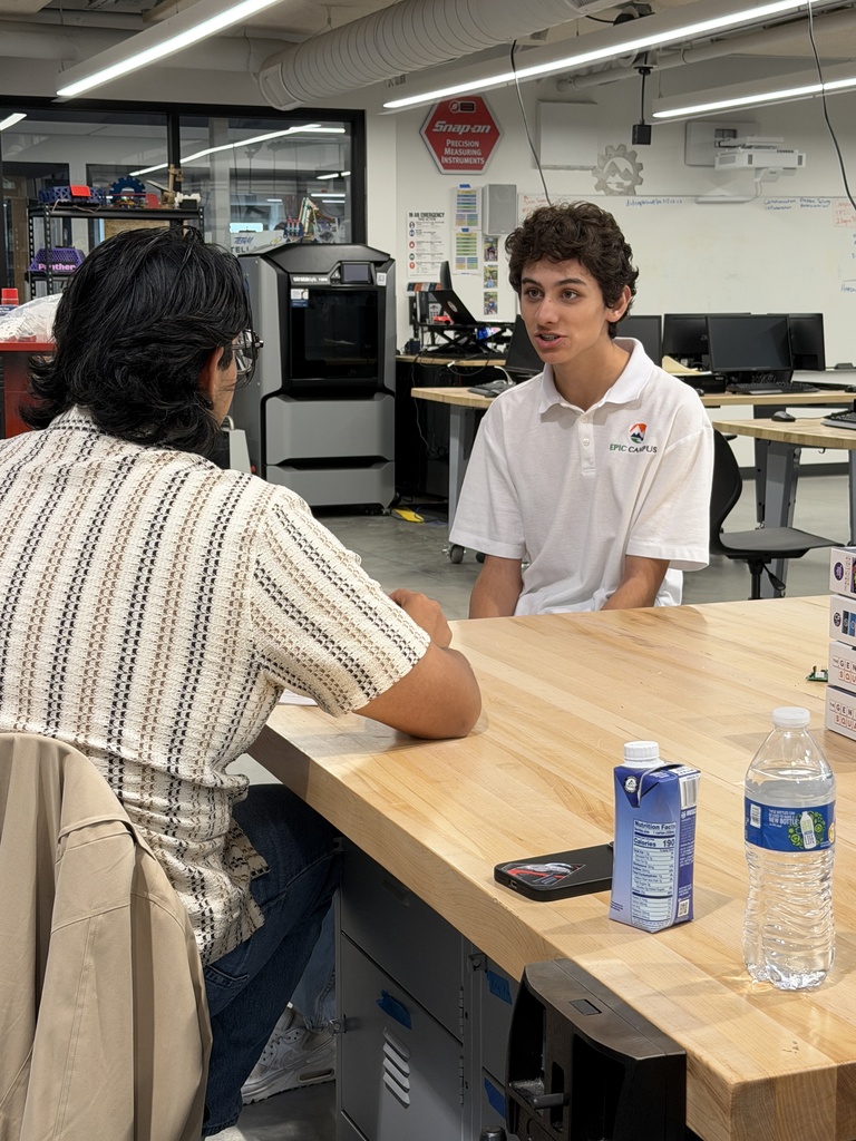 Two people sit at a desk in an office. A bottle, cellphone, and small box are on the desk.