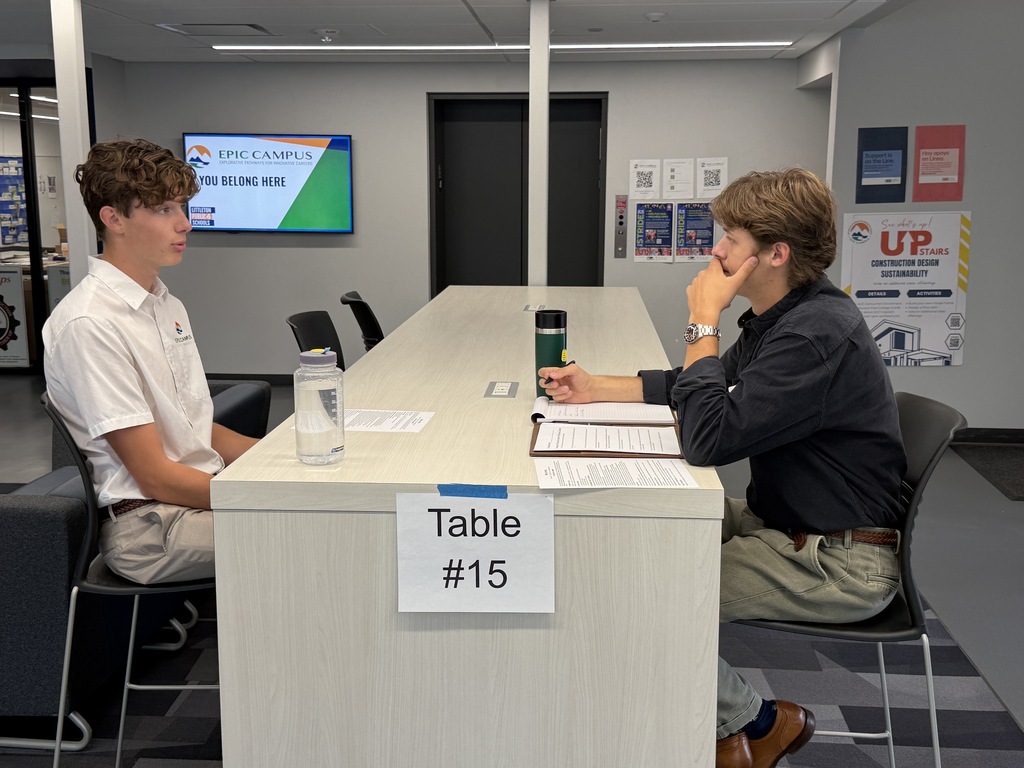 Two men sitting at a long table, one wearing a white shirt and the other in black. A table sign says "Table #15".